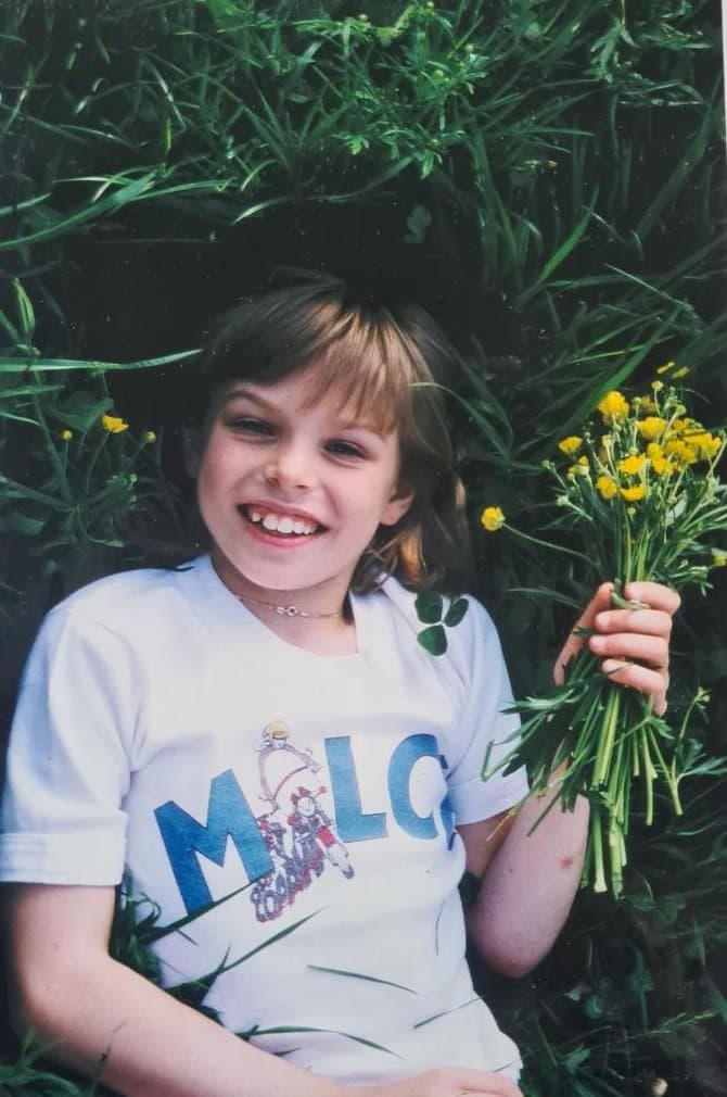 Ariane als Kind, lachend im Gras mit einem Strauß gelber Wiesenblumen