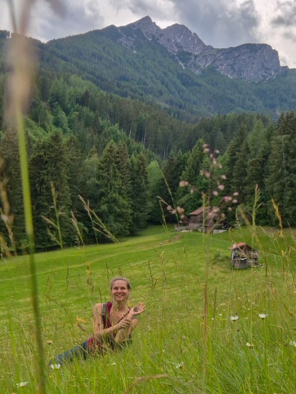 Ariane sitzt lachend in einer Bergwiese, hinter ihr Wald und Gipfel